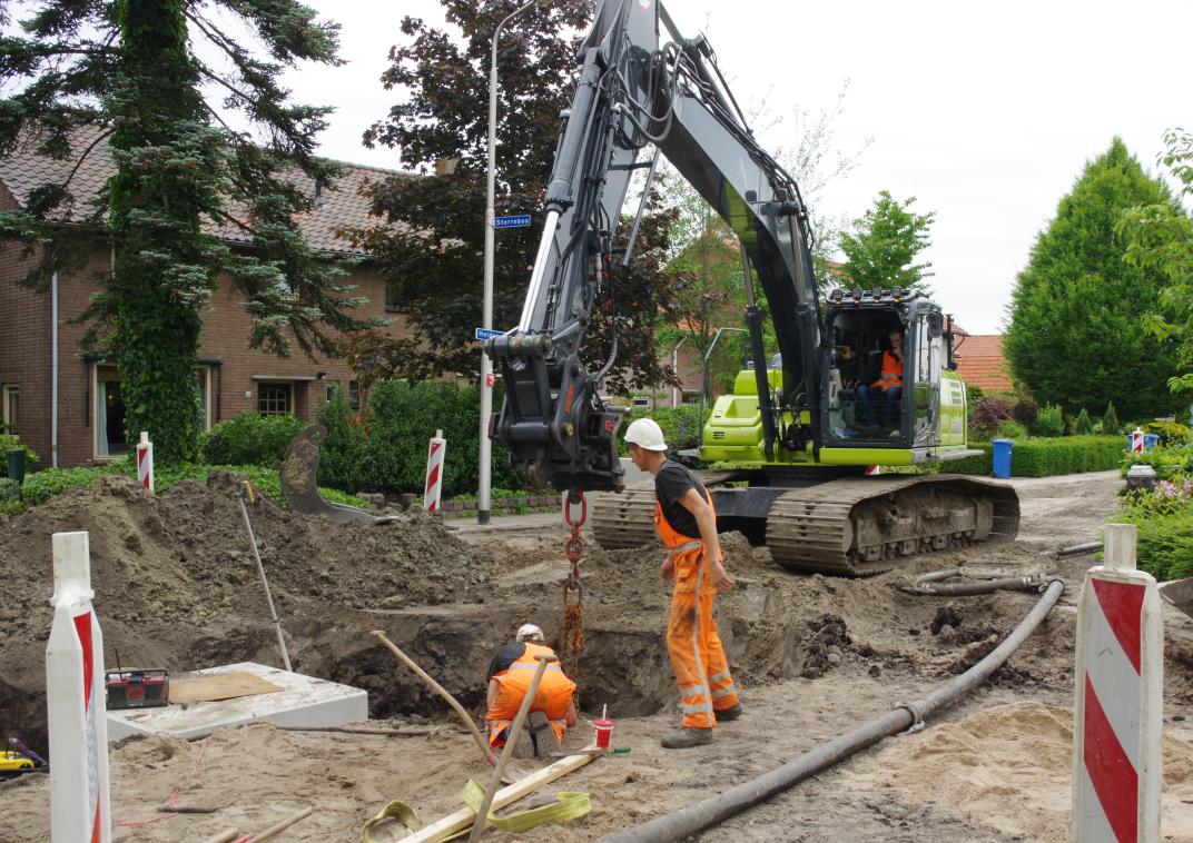 werkzaamheden in een straat in Hooglanderveen; een kraanwagenbediende haalt met zijn kraanwagen iets uit de opengebroken straat terwijl zijn collega's bij het gat in de weg staan
