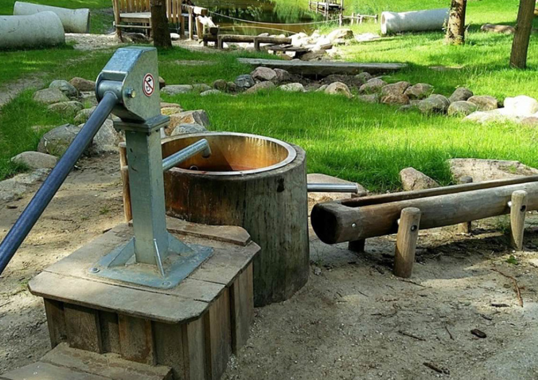 Een grote metalen waterpomp op een houten verhoging met daarnaast een ronde houten ton en een houten watergoot. Op de achtergrond is vers groen gras te zien.