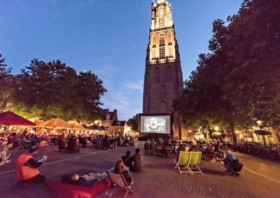 Een plein omringd door bomen en op het plein zitten liggen mensen te kijken naar een groot beeldscherm. Het is avond, lichten schijnen en op de achtergrond staat een hoge toren die van boven ook verlicht is.