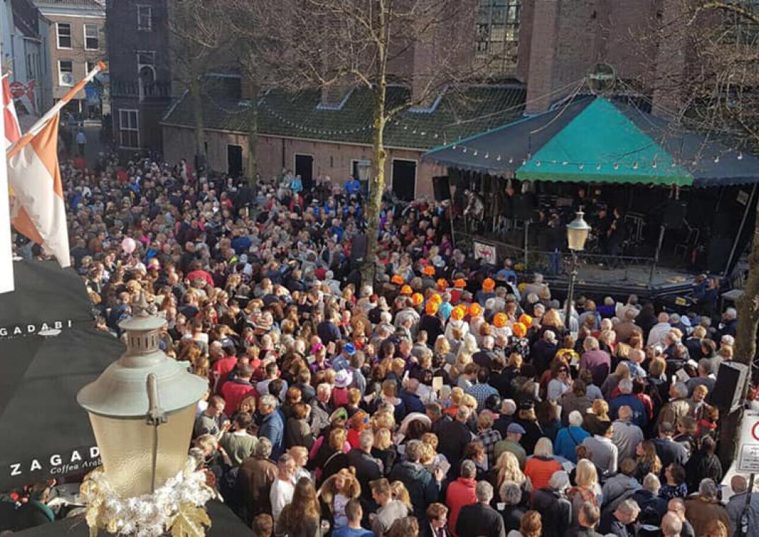Een grote mensen menigte staat voor een muziektent waar muzikanten in zitten