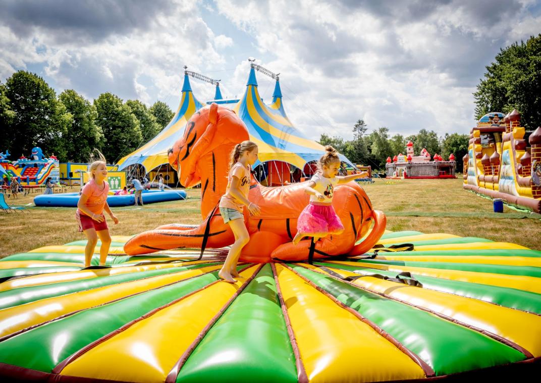 3 meisjes springen op een rond springkussen met in het midden een opblaasbare tijger. Op de achtergrond staan verschillende springkussens, opblaasbare stormbanen, een opblaaszwembad en in het midden staat een grote circustent 