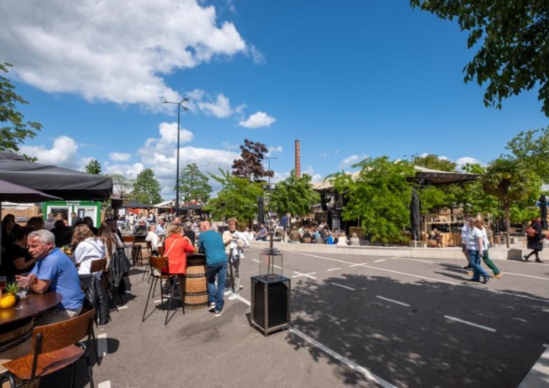 mensen zitten aan tafels op de achtergrond ligt achter bomen een podium