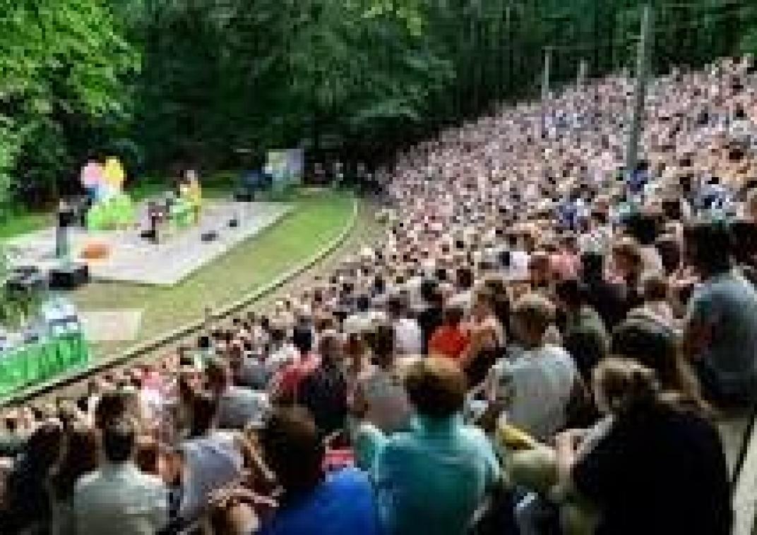 Een half ronde buitentribune is gevuld met rijen mensen voor de tribune is een podium met gras eromheen en daarachter staan allemaal bomen.