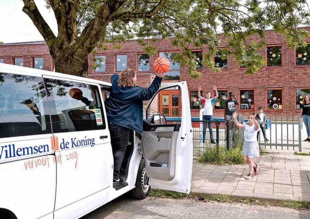 een witte bus met de tekst Willemsen de Koning staat geparkeerd. De portier staat open en in de deuropening staat een vrouw met een basketbal in haar hand die ze wil gooien naar een meisje