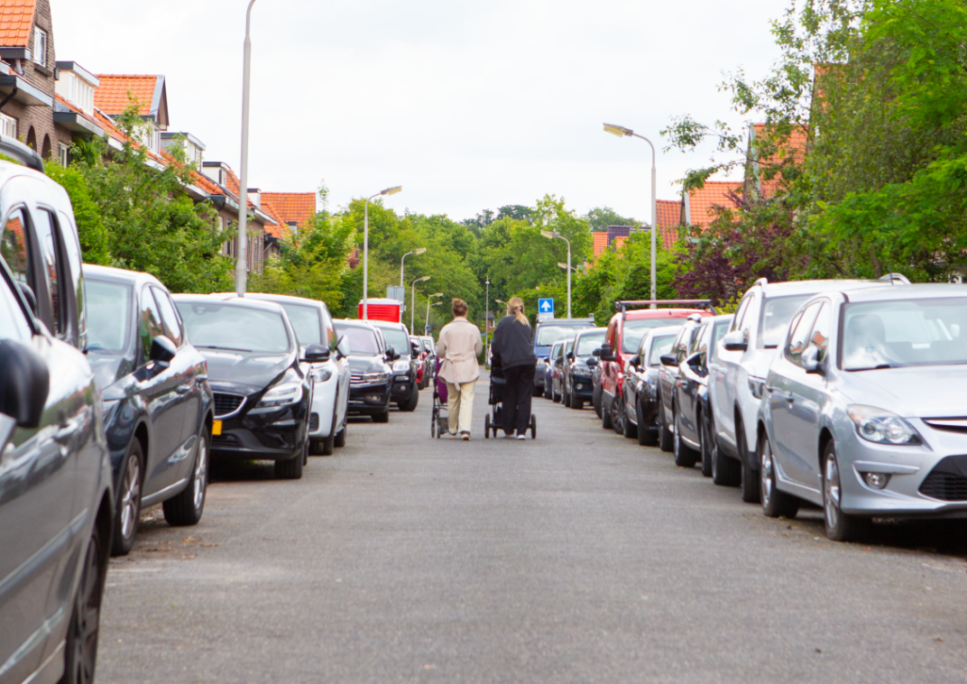 Een straat met aan allebei de kanten geparkeerde auto's en in het midden lopen twee vrouwen met een kinderwagen.