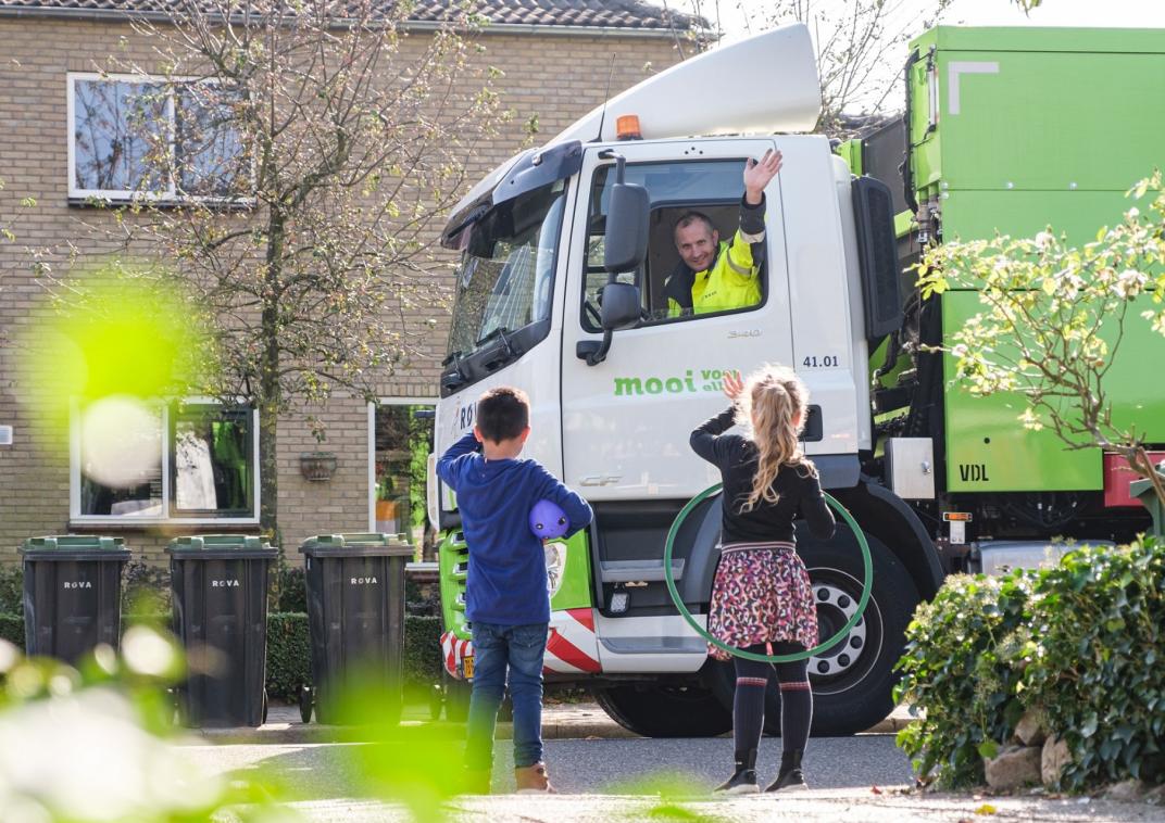 Een vrachtwagen van ROVA waarvan de chauffeur vriendelijk zwaait naar twee kinderen die terugzwaaien.