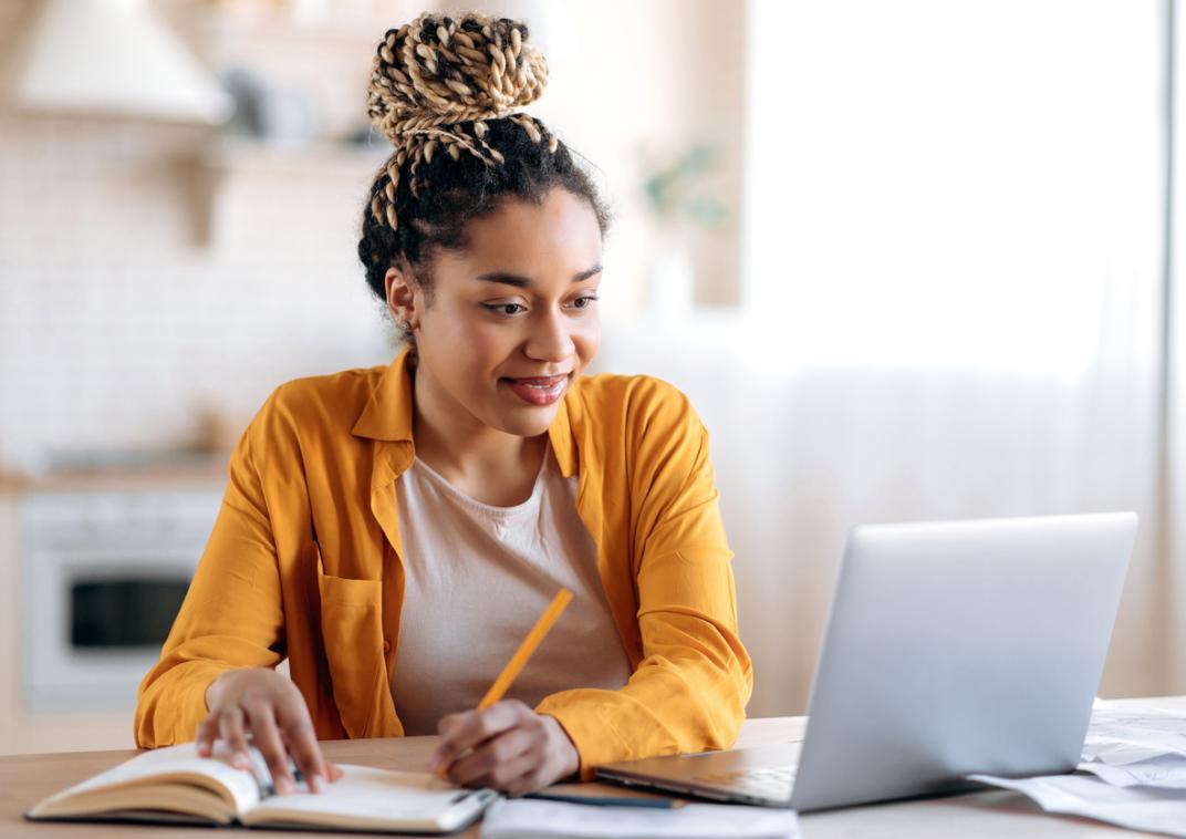 Een vrouw zit aan een tafel, kijkt naar een laptop en schrijft iets op in een notitieblok.
