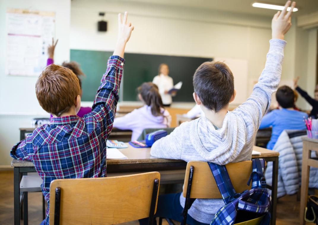 Kinderen zitten in een klaslokaal en kijken naar een leraar voor het schoolbord. De kinderen steken allemaal hun vinger in de lucht.