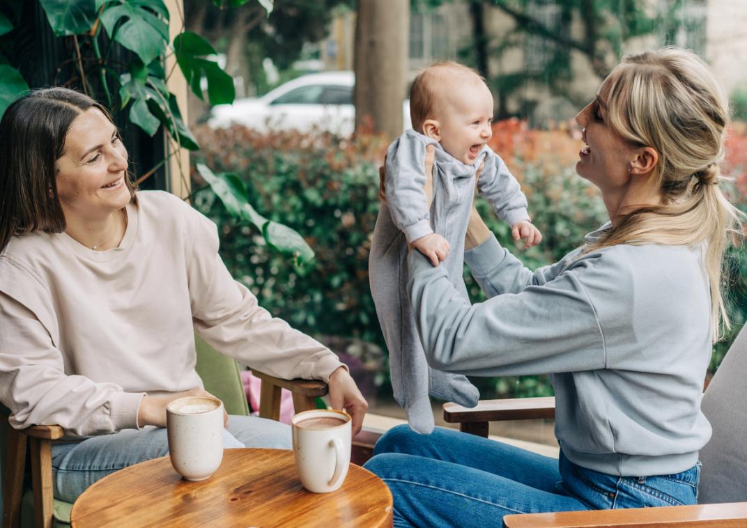 Twee vrouwen zitten op een terras met een baby en twee koppen koffie.