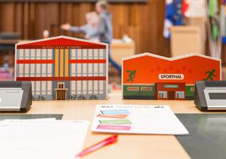 Kartonnen miniatuurversies van een middelbare school en een sporthal  op een tafel in de raadzaal