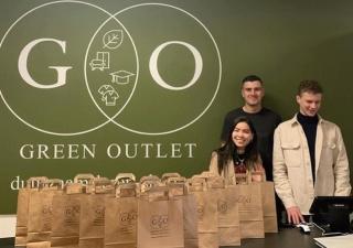 Drie jonge mensen staan achter een toonbank. Achter de mensen staat een groen scherm met daarop de tekst: GO Green Outlet