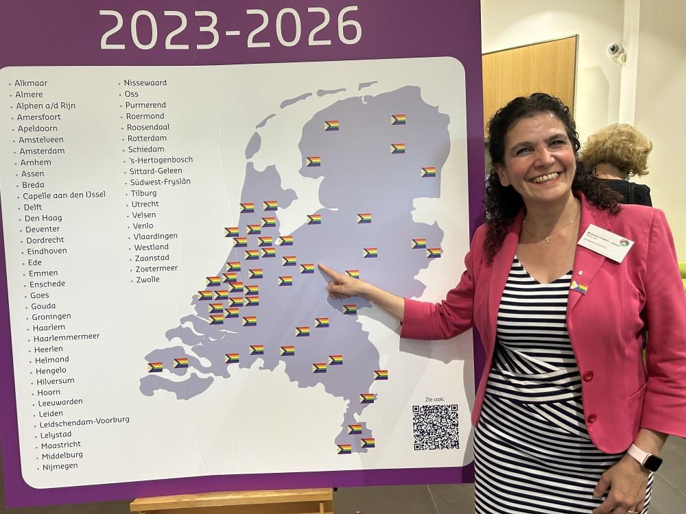 Foto van Wethouder Micheline Paffen-Zeenni bij een kaart van Nederland met daarop aangegeven de regenboogsteden