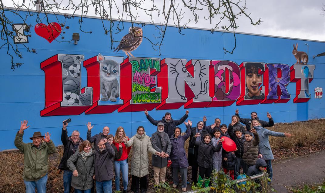 Een groepsfoto van mensen die voor een grote blauwe muur staan met daarop geschilderd in grote letters: Liendert