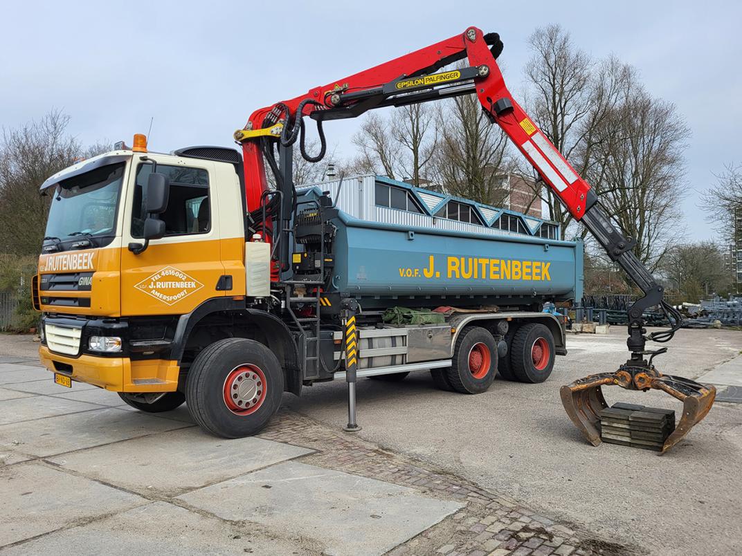 Een kraanwagen met bak waarvan de grijper op de grond staat met een stapel tegels tussen zijn geopende klauwen