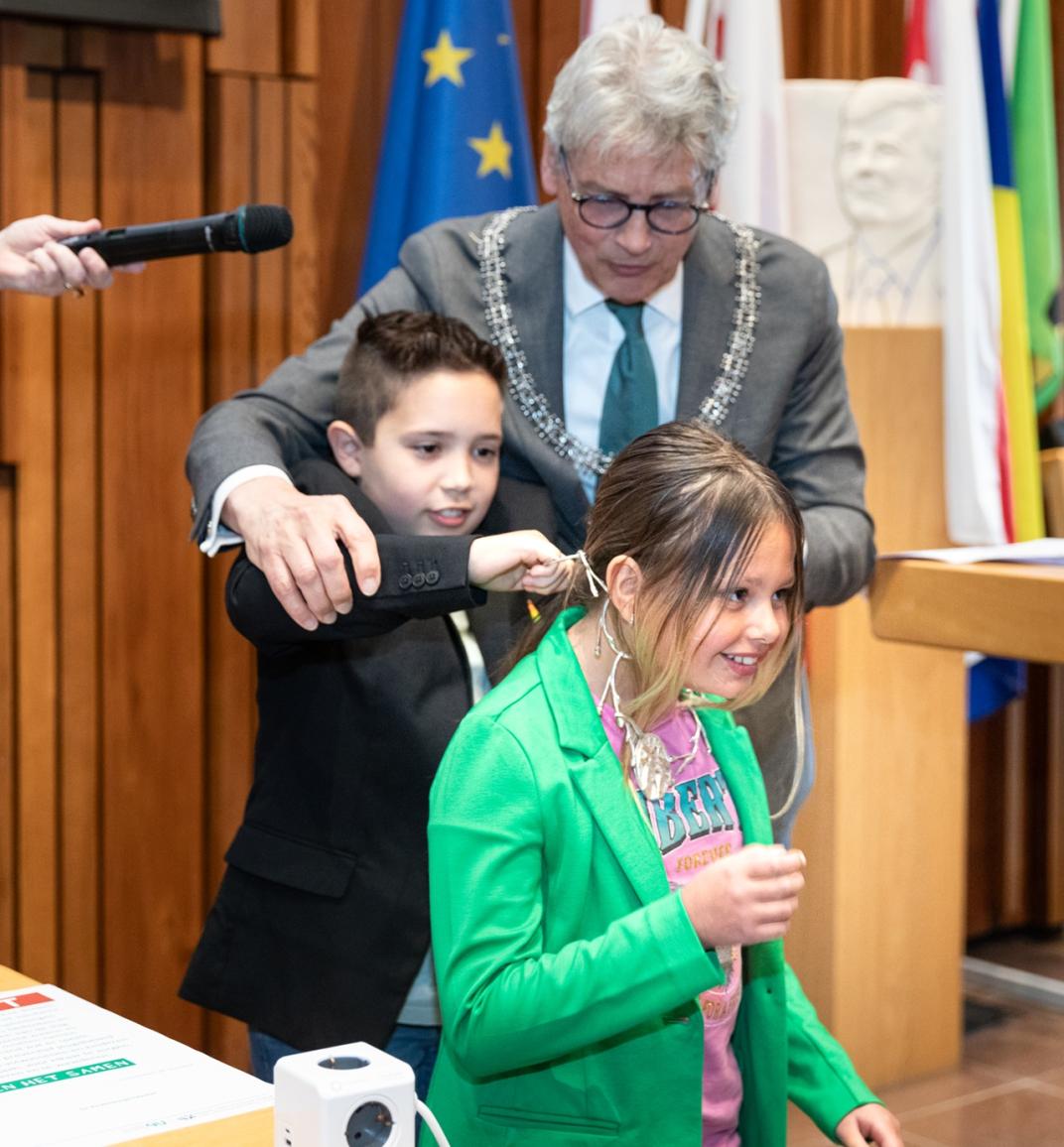 Oud-kinderburgemeester Aiden hangt de keten om bij de nieuwe kinderburgemeester Djennah, met op de achtergrond burgemeester Bolsius