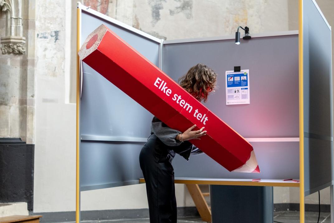 Een vrouw met een potlood zo groot als zijzelf vult in een stemhokje een stembiljet in