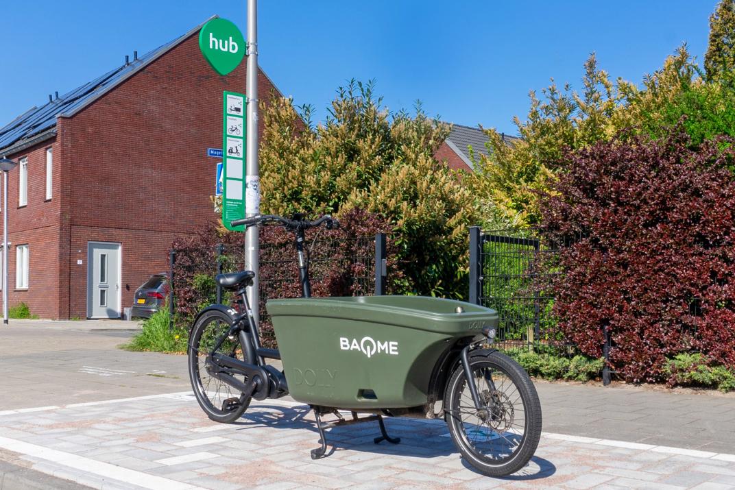 Deze foto toont een moderne bakfiets die geparkeerd staat op een speciaal ingerichte mobiliteitshub in een woonwijk. Op de voorgrond staat een donkergroene elektrische bakfiets met een grote laadbak. Op de laadbak staat de naam “BAQME”. De fiets staat op een verhard vak en rust op zijn standaard. Achter de fiets staat een paal met een groen “hub”-bord, waarop pictogrammen te zien zijn voor verschillende vormen van gedeelde mobiliteit, zoals fietsen en mogelijk andere vervoermiddelen.