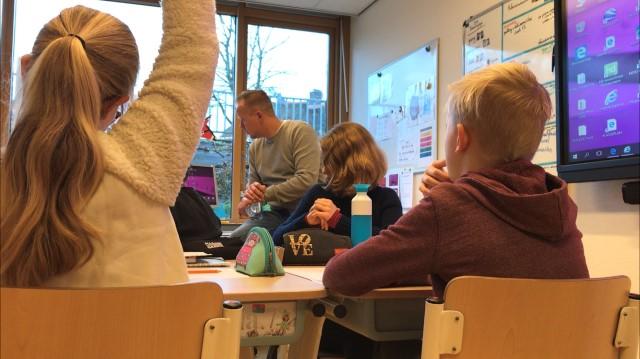 Foto van leerlingen in een klas; de leraar zit op zijn bureau en een meisje steekt haar hand op