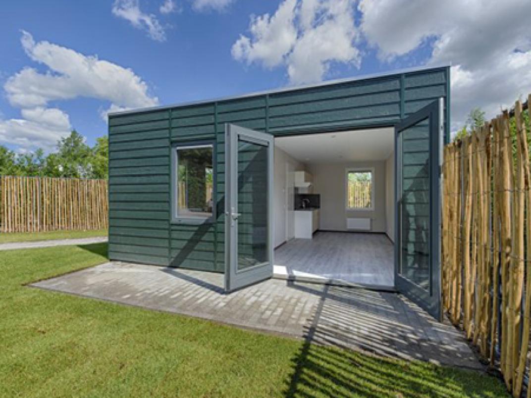 Skaeve Huse in Rotterdam; een klein groenblauw gebouw gelegen in een tuin.  De voorgrond is gevuld met weelderig groen gras, terwijl de achtergrond een uitgestrekte blauwe lucht laat zien. De tuin wordt gescheiden door een schutting gemaakt van lange takken.