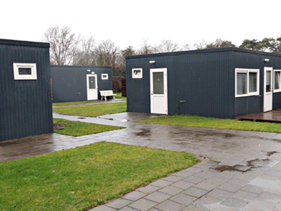 Skaeve Huse in Hilversum; een groep kleine gebouwen gelegen in een tuin. De dominante kleuren zijn wit en donkergrijs. De voorgrond is gevuld met weelderig groen gras, terwijl de achtergrond een uitgestrekte blauwe lucht laat zien. 